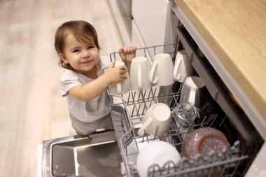 Little cute toddler girl helping to unload dishwasher. Funny little mommys helper in white kitchen at home. Healthy kid is doing household chores. Child indulges in the kitchen.