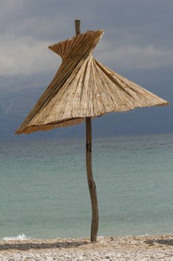 Sahilde yalnız bir kamış şemsiyesi, arka planda dağlar ve hava durumu istikrarsız ve fırtına mümkün, Baska, Krk adası, Hırvatistan