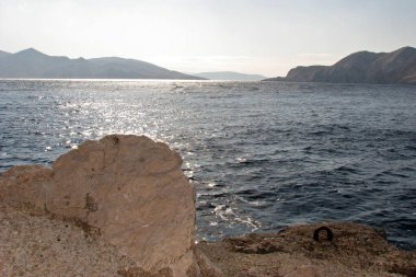 Gemi için demirleyen kayalık bir sahil ve güneş ışığında parıldayan deniz yüzeyine bakan bir manzara, adanın kayalık tepeleri, Baka, Hırvatistan 'ın Krk adası