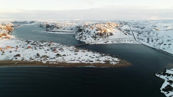 O drone voa sobre a vila piscatória na baía do Mar do Norte, a ponte conecta a ilha ao continente. O clima severo da península de Kola. costas cobertas de neve de Teriberka — Vídeo de Stock