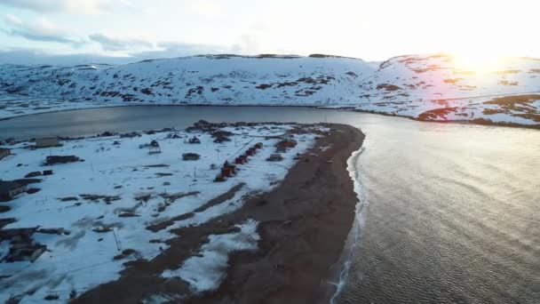 El dron vuela bajo sobre un pueblo pesquero en la bahía del Mar del Norte. El duro clima de la península de Kola en los rayos del sol poniente. En la costa nevada de Teriberka Metraje De Stock