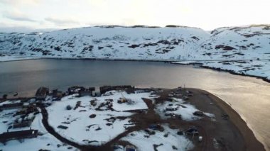 İnsansız hava aracı, karla kaplı uçurumlarla çevrili küçük bir kuzey köyünün üzerinden uçuyor. Sert kuzey iklimi. Teriberka köyü, Murmansk bölgesi.