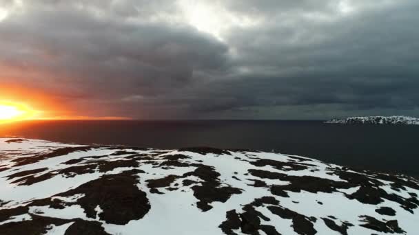 Um pôr-do-sol incrível sobre o oceano com nuvens de tempestade. O drone voa nas águas negras do oceano que lavam a costa rochosa da Escandinávia ou América do Norte. Oceano Ártico, rochas cobertas de neve — Vídeo de Stock