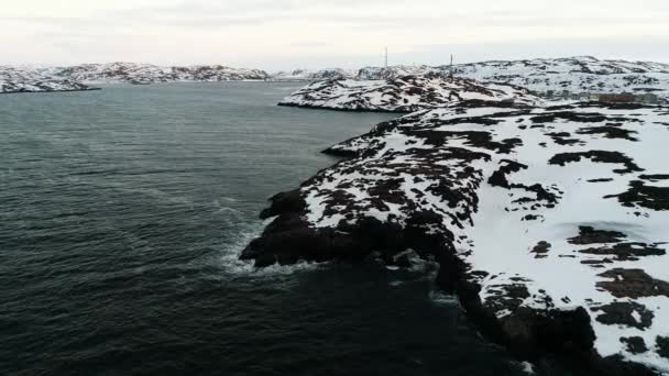 Dron létá v černých vodách oceánu, které omývají skalnatý břeh pokrytý sněhem. Arktický oceán Drsné podnebí poloostrova Kola. Zasněžené břehy Teriberky — Stock video