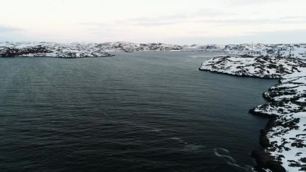 El dron se eleva por encima de las oscuras aguas del Océano Ártico cerca de la costa rocosa cubierta de nieve. El duro clima de la península de Kola Cubierta de nieve en las costas de Teriberka Un pueblo en la costa norte. Clip De Vídeo