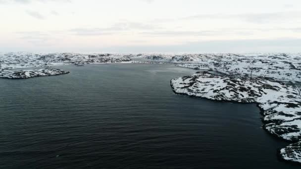 Dron letí v zátoce Severního ledového oceánu ke skalnatým břehům. Drsné podnebí poloostrova Kola. Krásné drone záběry tmavé vody a země pokryté sněhem — Stock video