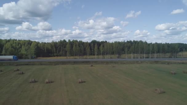 El dron vuela sobre el campo con fardos hacia el bosque donde hay una pista a lo largo de la cual los camiones se mueven con y sin remolque. La logística del transporte es el motor de la economía Clip De Vídeo