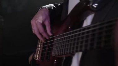 Gece elbisesi giymiş bir müzisyenin çaldığı bas gitarın yakın çekimi. Bir gece kulübündeki konserde bir müzik grubunun performansları. Rock grubu müzisyeni partnerleriyle bas gitar çalar.