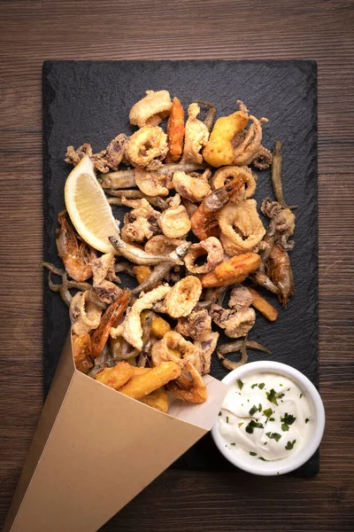 takeaway container with potatoes and fried squid. fried potatoes and squid on a slate board with mayonnaise and parsley.