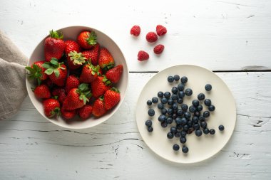 Beyaz ahşap arka planda bir kase çilek ve bir tabak yaban mersini. tepeden görünüm