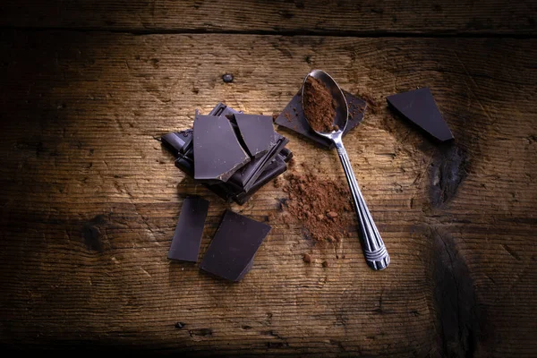 ココアとスプーンの近くに積み上げられたチョコレートの正方形 木製のテーブルの上 ダークチョコレートとココアパウダー — ストック写真