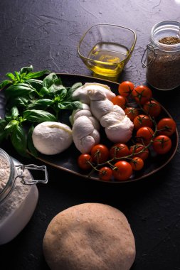 İçinde malzemeler olan pizza hamuru, mozarella, domates, fesleğen, un, koyu arkaplanda zeytinyağı.