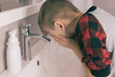 Küçük sarışın çocuk yüzünü banyoda yıkıyor.