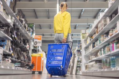 a girl with blond hair tied in a ponytail in jeans and a yellow sweater stands between the counters in a store with a blue plastic cart turned her back. High quality photo