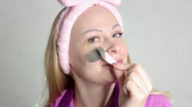 a cheerful girl of European appearance with blond hair on her head with a pink bandage,applies a mask on her face in gray color.behind a light background close-up of the face. High quality 4k footage