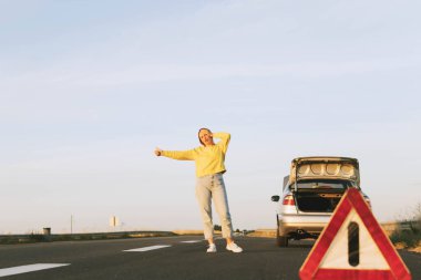in the foreground, a red-white emergency stop sign, in the background, a girl stands with her hand raised, stops the car for help on the road, upset. Car breakdown on the road. High quality photo