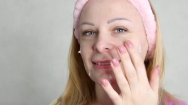 a smiling girl with a pink bandage on her head applies a face cream. A girl of European appearance with a tattoo on her left arm. The concept of a well-groomed and young face. High quality 4k footage