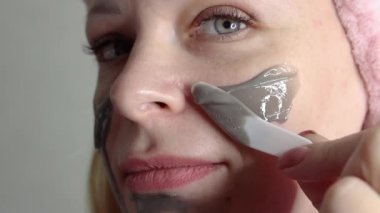  girl of European appearance with blond hair on her head with a pink bandage,applies a mask on her face in gray color.behind a light background close-up of the face,. High quality FullHD footage