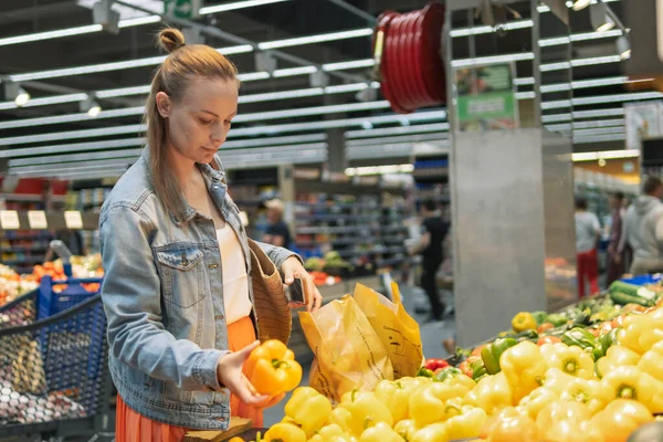 a girl of fair appearance, in a skirt and a denim jacket, stands near the counter with yellow salad pepper, puts it in a paper bag. The concept of healthy eating and vegetarianism. High quality photo