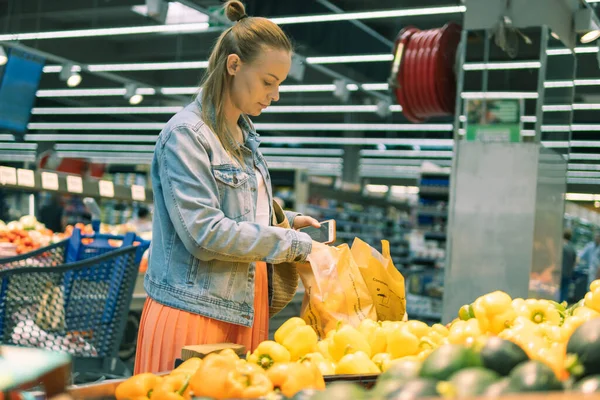 a girl of fair appearance, in a skirt and a denim jacket, stands near the counter with delta salad pepper, puts it in a paper bag. The concept of healthy eating and vegetarianism. High quality photo