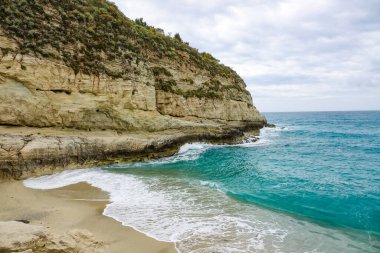 Turkuaz körfez, kumlu sahil, yeşil dağlar ve bitkiler, mavi gökyüzü, beyaz bulutlar arka plan, Capo Vaticano kaya platformu, Calabria, güney İtalya.