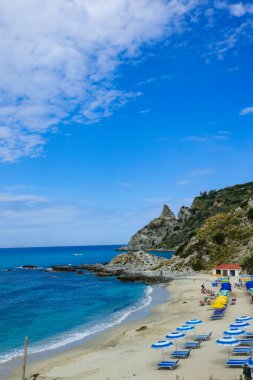 Turkuaz körfez, kumlu sahil, yeşil dağlar ve bitkiler, mavi gökyüzü, beyaz bulutlar arka plan, Capo Vaticano kaya platformu, Calabria, güney İtalya.