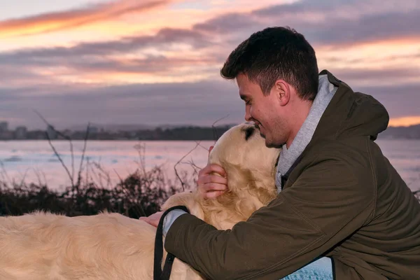 Genç adam gün batımında Golden Retriever köpeğine sarılıp oynuyor.