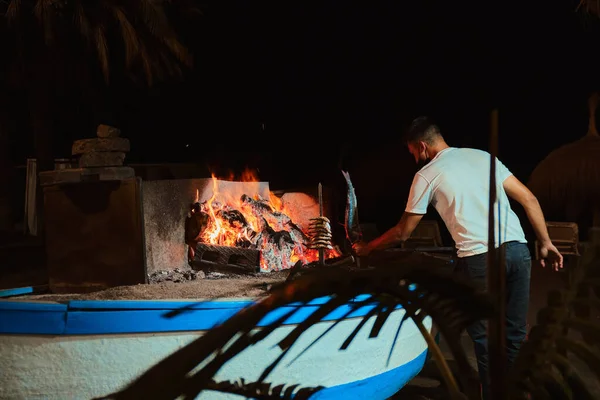 Malaga sahilinde bir teknede, genç bir garsonla barbekü. Geceleri sardalya broşürü pişiriyor.