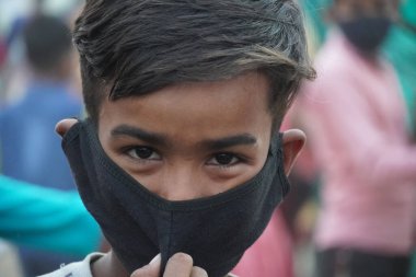 Bir çocuk maske takıyor ve kamera izliyor.