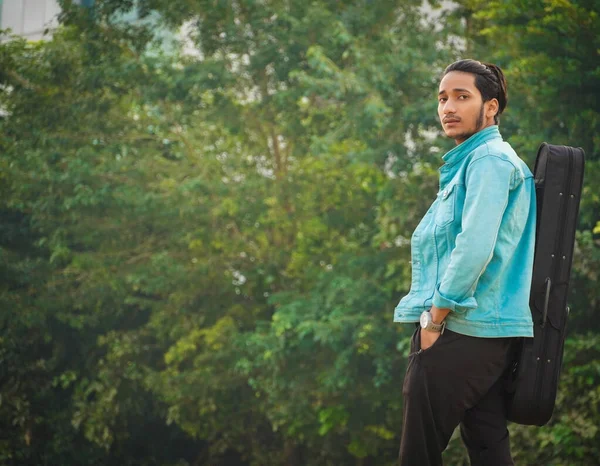 Yakışıklı hippi genç adam keman çantasıyla ayakta mutlu ve havalı bir gülümsemeyle gülümsüyor..