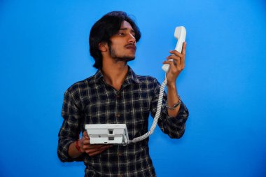 Yakışıklı genç adam mavi arka planda telefonla konuşuyor.