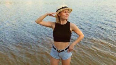 Şapkalı ve kısa kot şortlu genç bir kadın dalgalarla birlikte suyun kenarında duruyor. Yazın deniz kenarında rahatla. Tatilde dinlenin. Suyun üstünde bir kadının portresi.