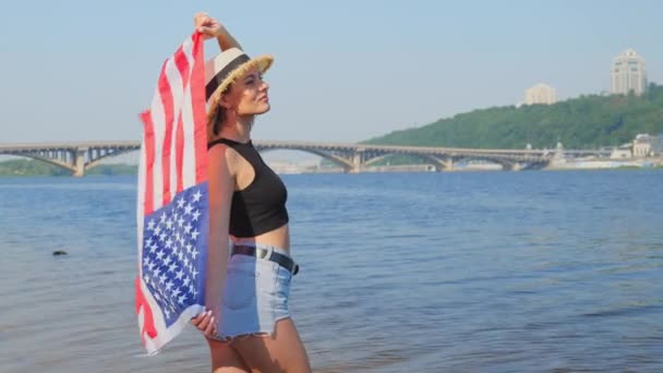 帽子とショートパンツの若い女性は 川と橋の近くで彼女の手に米国の国旗を持っています スローモーション 水の近くの女性 独立記念日 — ストック動画