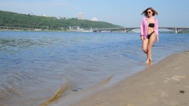Siyah mayosu ve pembe gömleği olan genç bir kadın sahile doğru yürüyor. Yavaş çekim gevşemesi. Yaz tatili