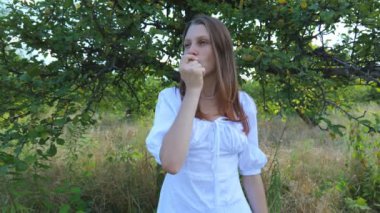 Beyaz elbiseli genç bir kadın bahçede yeşil bir elma yiyor. Yazın dinlen.