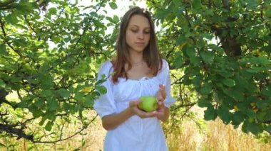 A young woman in a white dress stands among the trees holding green apples in her hands. Summer holidays in the botanical garden