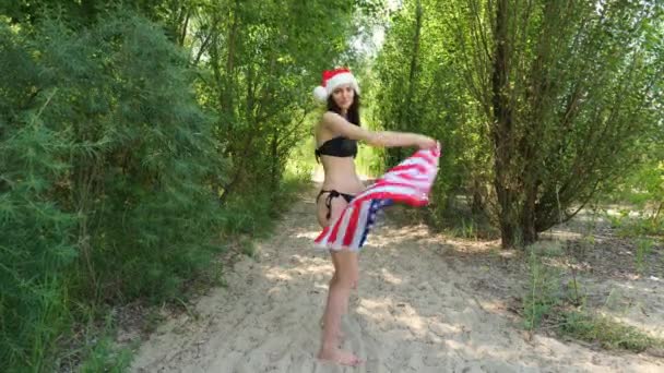 一个身穿黑色泳衣的女孩在树间沙滩上散步的后视镜 布鲁内特女人拿着美国国旗独立日 假日休息 — 图库视频影像