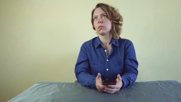 デニムシャツを着た40歳の女性が灰色のテーブルに座っている 大人の女性は彼女の手に携帯電話を考え 保持します スローモーション4K — ストック動画