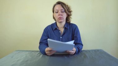 Kırk yaşında kot gömlekli bir kadın gri bir masada otururken elinde beyaz bir kağıt tutuyor. Yavaş çekim portresi