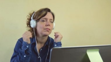 Kırk yaşında, mavi gömlekli, büyük kulaklıklı bir kadın dizüstü bilgisayarın başında oturuyor. Müzik dinleyen yetişkin bir kadın.