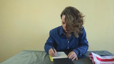 Kırk yaşında kot gömlekli bir kadın masada otururken kağıt bir defterde kalemle yazar. Yetişkin bir kadının portresi