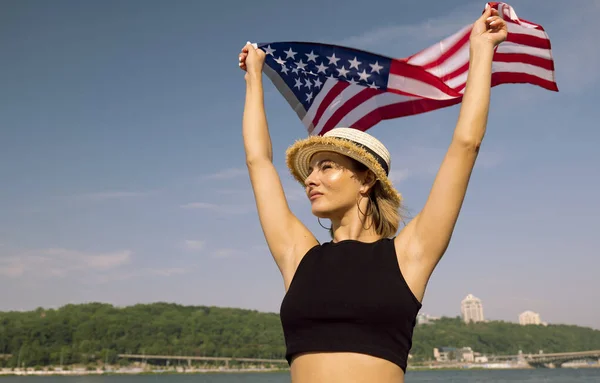 Şehirde nehir kenarında Amerikan bayrağı taşıyan şapkalı bir kadın. 4 Temmuz Bağımsızlık Günü. Siyah askılı Amerikalı bir kadının portresi.
