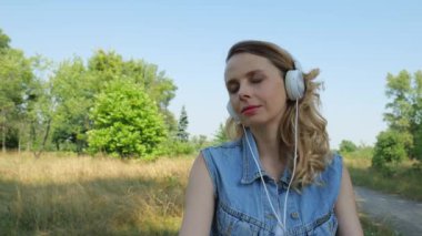 Kırk yaşında kot elbiseli yetişkin bir kadın büyük beyaz kulaklıklarla müzik dinliyor. Doğada sarışın bir kadının portresi. yavaş çekim