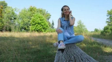 Kot elbiseli bir kadının ahşap bir bankta oturduğunu görmek. Bir kadın, doğada otururken beyaz kablo kulaklıkla müzik dinler. Yavaş çekim portresi