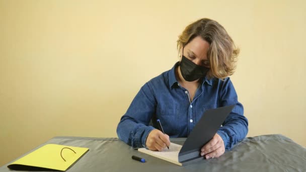 デニムシャツを着た40歳の女性が紙のノートに書かれたテーブルに座っている 黒マスクの大人の女性でテーブルに座って — ストック動画