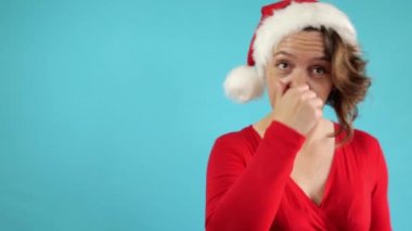 Kırk yaşında kırmızı kazaklı bir kadın burnuna dokunuyor. Noel şapkalı yetişkin bir kadının portresi. Mavi arka planda Amerikalı bir kadın. Yavaş çekim 4k