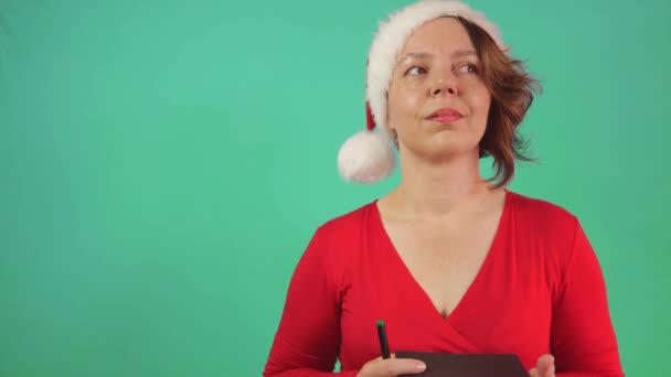 赤いセーターを着た40歳の女性の肖像画と 緑の背景に紙のノートが付いた新年の帽子 スローモーション 新年の願い メリークリスマス — ストック動画