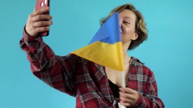 Ekose gömlekli yetişkin bir Amerikalı kadın mavi arka planda Ukrayna bayrağıyla selfie çekiyor. Yavaş çekim portresi. 40 yaşında güzel bir kadın.
