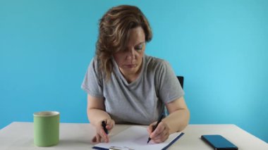 Gri tişörtlü yetişkin bir kadın mavi arka planda kağıtları olan bir masada çalışıyor. Bir kadın masada siyad yazıyor. yavaş çekim