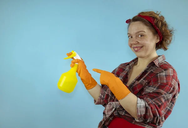 A cheerful pin-up woman in a plaid shirt holds a yellow spray bottle in her hands on a blue background. Woman cleaning on a blue background. woman in a shirt. Pin up style